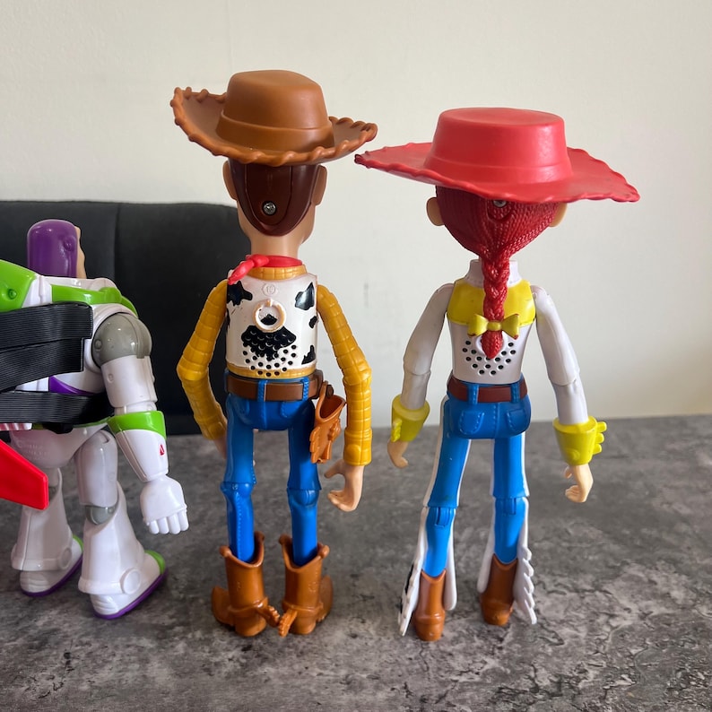 May include: Three plastic Toy Story character figures. Woody and Jessie are in the center, wearing cowboy hats, vests, and boots. Buzz Lightyear is on the left, in a green and purple spacesuit.