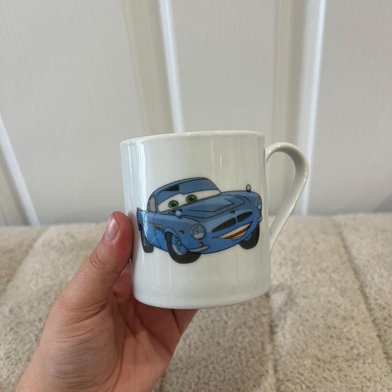 May include: A white ceramic mug featuring a graphic illustration of a light blue anthropomorphic car with large green eyes, a wide smiling mouth, and black tyres. The mug has a smooth, glossy finish and a curved handle on the right side.