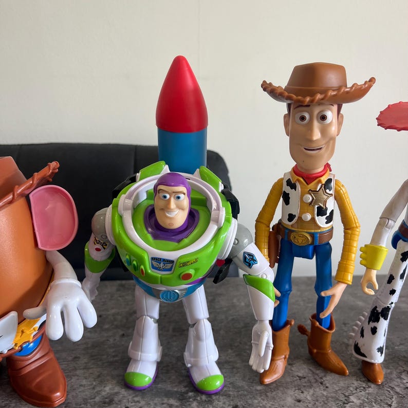 May include: A selection of Toy Story character figures. Buzz Lightyear in a green and white spacesuit with a red and blue rocket. Woody in a cowboy outfit with a brown hat. Other characters are also visible.