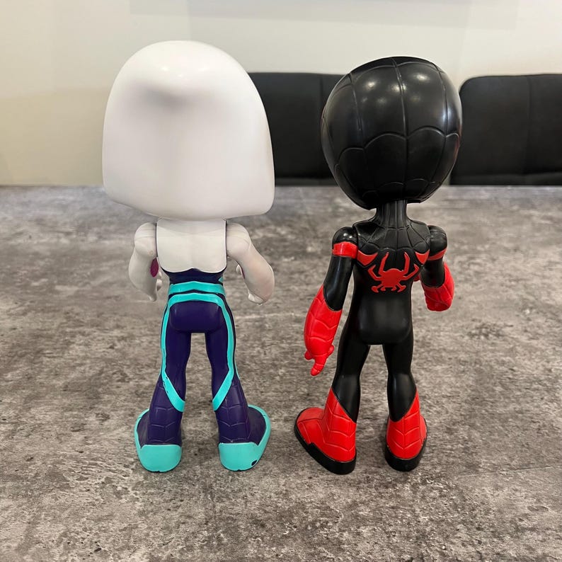 May include: Two small action figures, one white with teal accents and one black with red accents. The white figure has a large head and is wearing a teal and purple outfit. The black figure has a large head and is wearing a red and black outfit with a spider symbol on the chest.