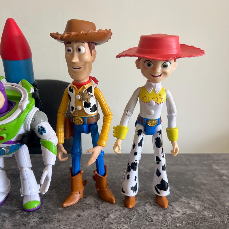 May include: Toy Story character figures of Woody, Jessie, and Buzz Lightyear. Woody is in a cowboy hat, vest, and boots. Jessie has a red hat and cow-print trousers. Buzz Lightyear is in a green and purple spacesuit.