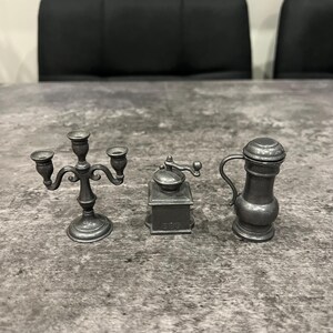 May include: Three miniature pewter items: a three-arm candelabra, a coffee grinder, and a pitcher with a lid. The items are arranged on a gray surface. The candelabra has a decorative, curved design. The coffee grinder has a crank handle.