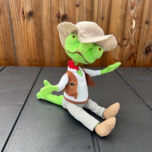 May include: A plush green lizard doll wearing a tan cowboy hat, a white shirt, a brown vest with a star badge, and gray pants. The doll has large yellow eyes and is posed sitting down with its arms outstretched.
