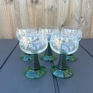 May include: A set of five clear glass wine glasses with a grape vine design etched on the bowl. The glasses have a green base and a black, ribbed stem. The glasses are arranged on a dark surface.