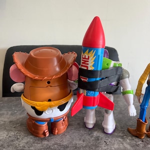 May include: Toy Story character figures including Mr. Potato Head wearing a cowboy hat, Buzz Lightyear with a rocket, and Woody in a cowboy outfit. The rocket is blue, red, and yellow with the word "JNE" on it.