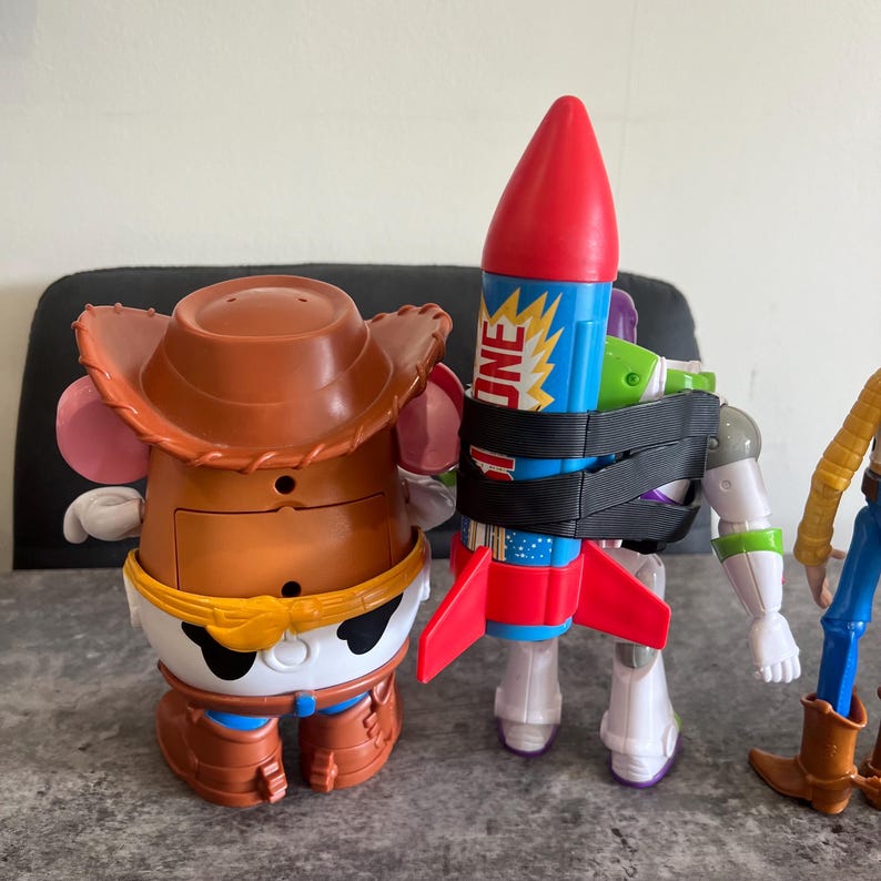 May include: Toy Story character figures including Mr. Potato Head wearing a cowboy hat, Buzz Lightyear with a rocket, and Woody in a cowboy outfit. The rocket is blue, red, and yellow with the word "JNE" on it.
