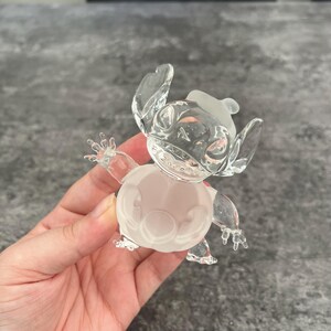 May include: A clear glass figurine of Stitch from the Disney movie Lilo & Stitch. The figurine is in a standing pose with its arms outstretched. The figurine is frosted white and has a clear glass body.