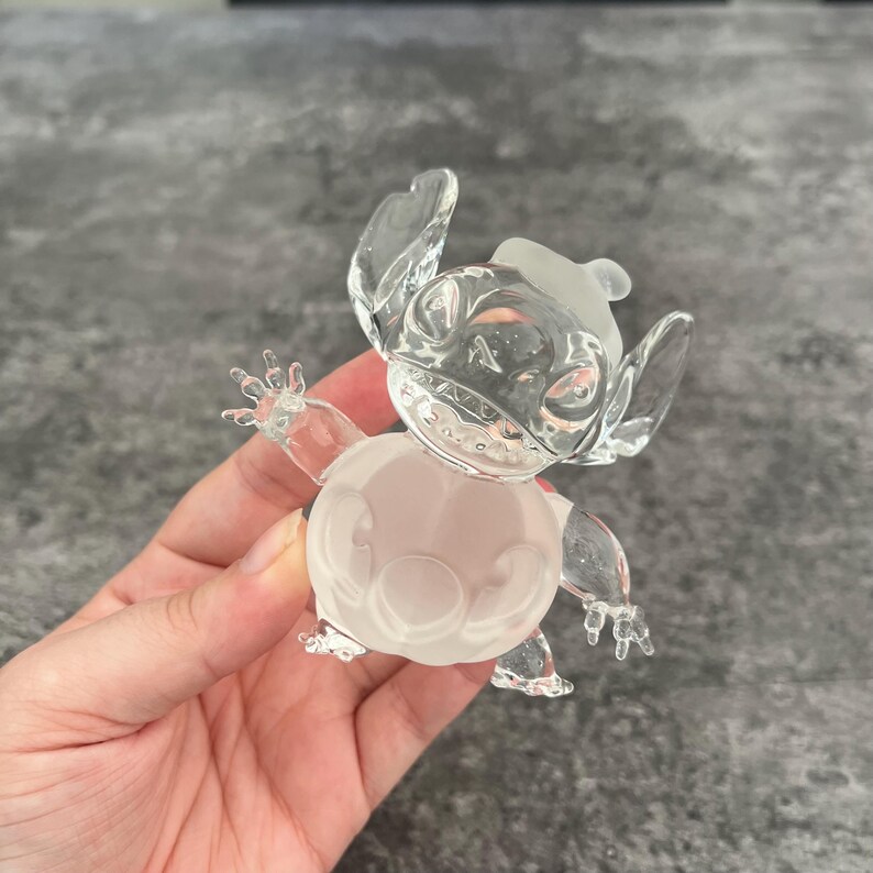 May include: A clear glass figurine of Stitch from the Disney movie Lilo & Stitch. The figurine is in a standing pose with its arms outstretched. The figurine is frosted white and has a clear glass body.