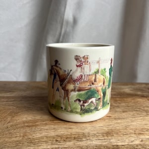 May include: Cream-colored ceramic mug with a vintage equestrian scene. The illustration depicts two children riding a horse, a dog, and figures in period clothing. The mug has a slightly flared shape and a smooth surface.