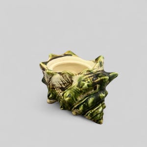May include: A ceramic seashell-shaped container with a glossy green glaze. The container has a cream-colored interior and a textured exterior, with a spiral design and pointed edges. The decorative piece is suitable for holding small items.