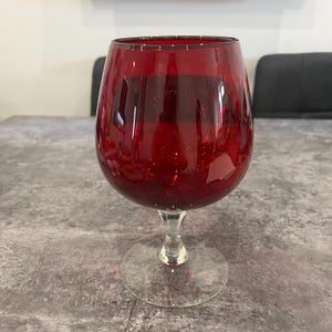 May include: A large, ruby red wine glass with a clear stem and base. The glass has a rounded bowl and is filled with a dark red liquid. The glass is on a grey surface.