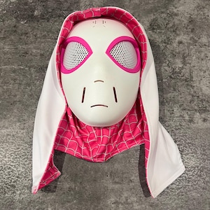May include: A white Spider-Gwen mask with pink detailing and mesh eye coverings. The mask is framed by a white hood with a pink spiderweb pattern lining. The mask has a stylized, neutral expression.