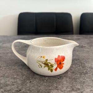 May include: Cream-colored ceramic creamer with a handle and spout. The creamer features a floral design with red and yellow flowers and green leaves. The creamer has a speckled finish and a gold rim.