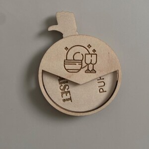 May include: A wooden round box with a hinged lid. The lid has a cutout of a hand pointing to the right. The box has a design of a plate and two glasses on the lid. The box is labeled "Purist" on the right side and "Asset" on the left side.