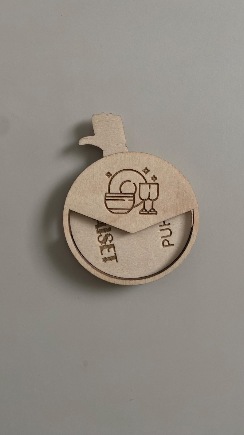 May include: A wooden round box with a hinged lid. The lid has a cutout of a hand pointing to the right. The box has a design of a plate and two glasses on the lid. The box is labeled "Purist" on the right side and "Asset" on the left side.
