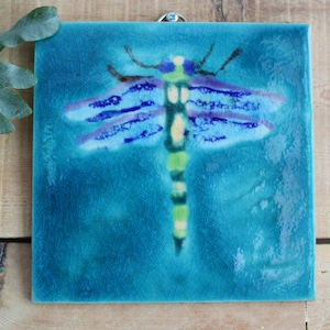 Pode incluir: Um azulejo de cerâmica azul com uma libélula pintada em tons de azul, verde e amarelo. A libélula tem um corpo longo e fino e asas delicadas.