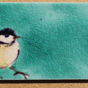 May include: A rectangular ceramic tile with a bird illustration. The bird has a black head, white face, yellow chest, and brown wings. The background is a textured teal colour.