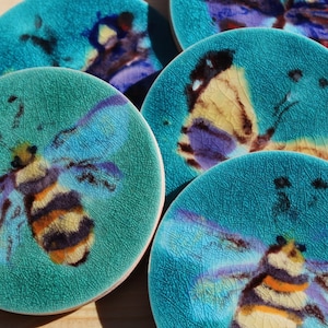 May include: Set of four ceramic coasters featuring a blue-green background with painted images of bees and butterflies. The coasters have a textured, crackled glaze.