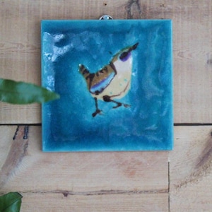 May include: A ceramic tile with a blue background and a painted image of a small bird with brown, yellow and blue feathers.
