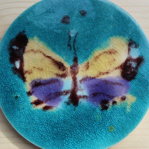 May include: A turquoise ceramic plate featuring a butterfly design. The butterfly is painted in shades of yellow, purple, and brown.