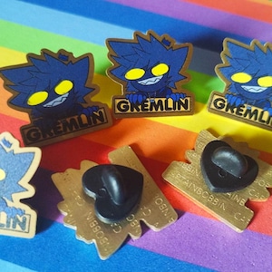 May include: A collection of enamel pins. Each pin features a blue gremlin character with yellow eyes and the word "GREMLIN". The pins are set against a rainbow-coloured background.