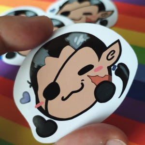 May include: A sticker with a cartoon character design. The character has a black eye patch, black ear, and a smiling expression. The character has light brown skin, black hair, and a pink blush. A small blue heart is visible.