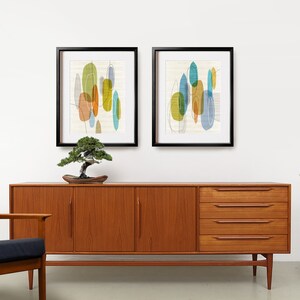 K&ouml;nnte beinhalten: Zwei gerahmte abstrakte Kunstdrucke mit geometrischen Formen in Blaut&ouml;nen, Gr&uuml;n, Gelb und Orange. Die Drucke h&auml;ngen an einer wei&szlig;en Wand &uuml;ber einem Sideboard aus Teakholz im Stil der Jahrhundertmitte.