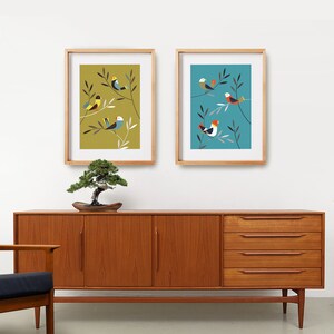 May include: Two framed prints featuring colourful bird illustrations on a light blue and a mustard yellow background. The prints are hanging on a white wall above a wooden dresser.