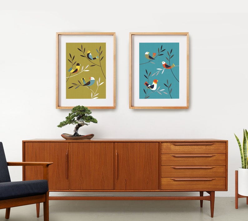 May include: Two framed prints featuring colourful bird illustrations on a light blue and a mustard yellow background. The prints are hanging on a white wall above a wooden dresser.