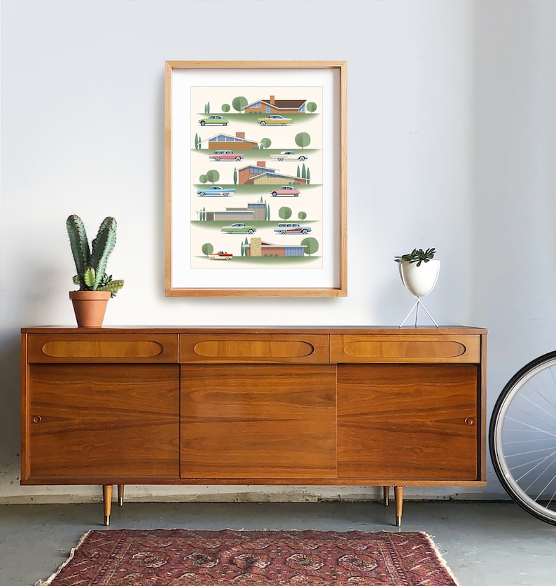 May include: A framed print of a mid-century modern illustration featuring a row of houses with cars in front of them. The houses are in various shades of orange, yellow, and brown. The cars are in shades of green, blue, red, and white. The background is a light cream color with green trees.