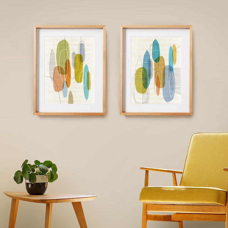 K&ouml;nnte beinhalten: Zwei gerahmte abstrakte Kunstdrucke im Mid-Century-Modern-Stil. Die Drucke zeigen bunte geometrische Formen und Linien auf wei&szlig;em Hintergrund. Die Rahmen sind in hellem Holz gehalten.