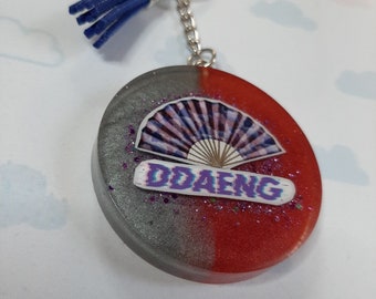 May include: A silver and red resin keychain with a blue tassel. The keychain features a decorative fan with a purple and white design and the word "DDAENG" in purple and white letters.
