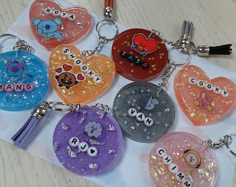 May include: Seven colorful resin keychains with different characters and names. The keychains are in the shape of hearts and circles. The characters include Koya, Shooky, Tata, Cooky, Van, RJ, and Chimmy.