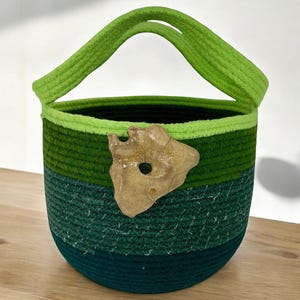 May include: A round, green and blue rope basket with a handle. The basket has a decorative tan-colored rock attached to the side. The basket is made of tightly woven rope and is suitable for storage.