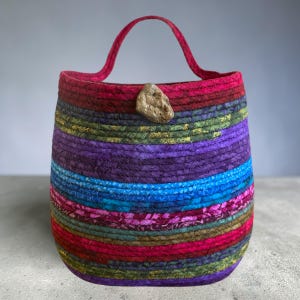 May include: A colorful, handmade tote bag with a rounded shape and a single handle. The bag is constructed from horizontal bands of fabric in various colors, including red, purple, blue, and green. A natural-colored stone adorns the side.