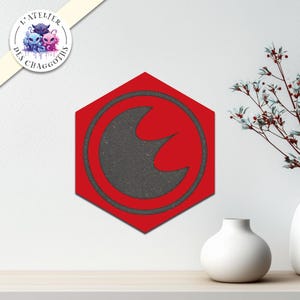 Könnte beinhalten: Rote, sechseckige Wanddekoration mit einem dunkelgrauen Emblem in einem Kreis. Das Emblem hat ein stilisiertes, geschwungenes Design. Die Dekoration befindet sich an einer weißen Wand, mit einer weißen Vase und einem dekorativen Zweig im Hintergrund.