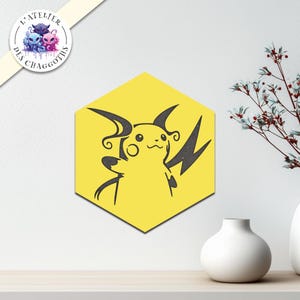 Peut inclure: Une œuvre d'art murale jaune en forme d'hexagone représentant une silhouette noire de Pikachu. L'œuvre est placée sur un mur blanc, avec un petit vase blanc et des branches décoratives en arrière-plan. L'œuvre est de "L'ATELIER DES CHAGGOTHS".