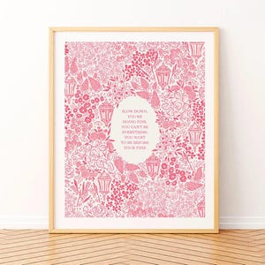 以下が含まれることがあります： ピンクの花柄の背景と、中央に「SLOW DOWN, YOU'RE DOING FINE, YOU CAN'T BE EVERYTHING YOU WANT TO BE BEFORE YOUR TIME」というテキストが入った楕円形が特徴の額入りプリント。 フレームは明るい木の色です。