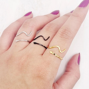 May include: A hand wearing four different wavy rings. The rings are silver, black, gold, and silver. The rings are all different sizes and shapes.