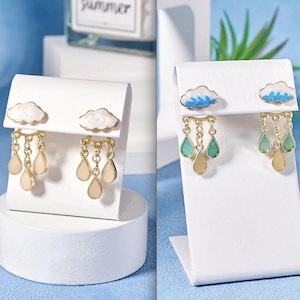 Peut inclure: Boucles d'oreilles en forme de nuage avec des breloques en forme de goutte. Les boucles d'oreilles présentent des nuages blancs avec des détails bleus et des chaînes dorées. Les gouttes sont beiges et vertes. Présentées sur des présentoirs blancs.