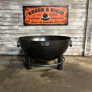 May include: A black metal fire pit with a round bowl and a ring stand. The fire pit is labeled "Rough & Rigid Custom Fire Pits Built in Kansas City".