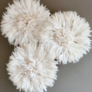 Op de afbeelding: Drie witte veren wanddecoraties, elk met een ronde vorm en een pluche textuur. De stukken zijn in een cluster op een grijze muur gerangschikt.