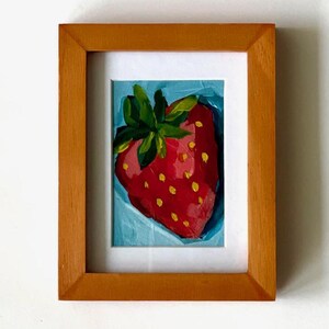 May include: A framed painting of a single red strawberry with yellow seeds and green leaves on a blue background.