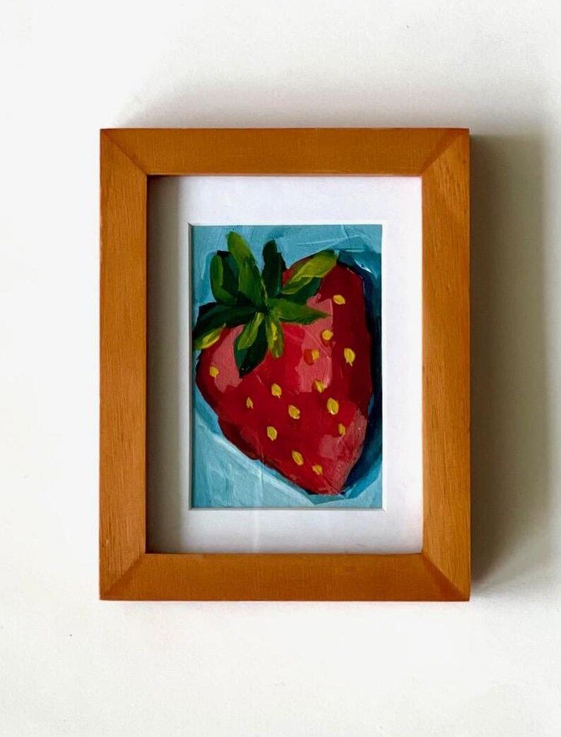 May include: A framed painting of a single red strawberry with yellow seeds and green leaves on a blue background.