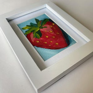 May include: A framed painting of a red strawberry with green leaves on a blue background.