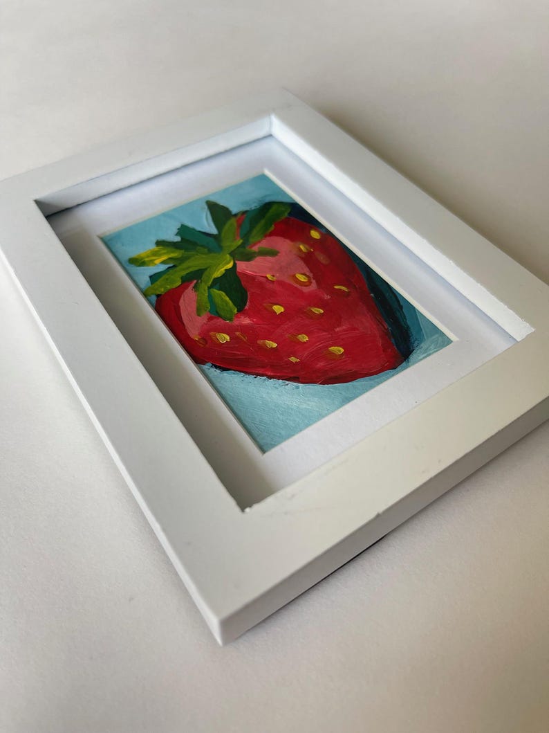 May include: A framed painting of a red strawberry with green leaves on a blue background.