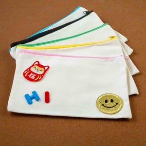 Peut inclure: Pochettes zippées en toile blanche avec des fermetures éclair colorées et des écussons décoratifs. Les écussons comprennent une poupée Daruma rouge, le mot "HI" et un smiley doré à paillettes. Les fermetures éclair sont noires, vertes, jaunes et bleues.