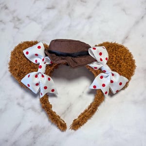 May include: Brown teddy bear ear headband with a small brown hat and white bows with red polka dots. The headband is a fun accessory for themed events or everyday wear.