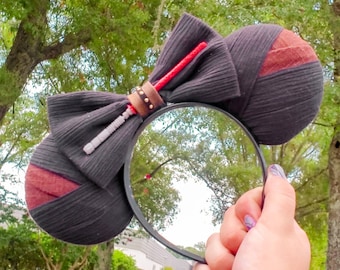 May include: Black and red themed mouse ears with a bow and lightsaber accents. The ears are black with red fabric accents. A red lightsaber and a gray lightsaber hilt are attached to the bow. The headband is black.