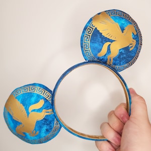 Può includere: Fascia blu e oro con due accenti auricolari circolari. Ogni orecchio presenta un design di Pegaso dorato all'interno di un bordo dorato a chiave greca su sfondo blu. La fascia è blu con interno dorato.